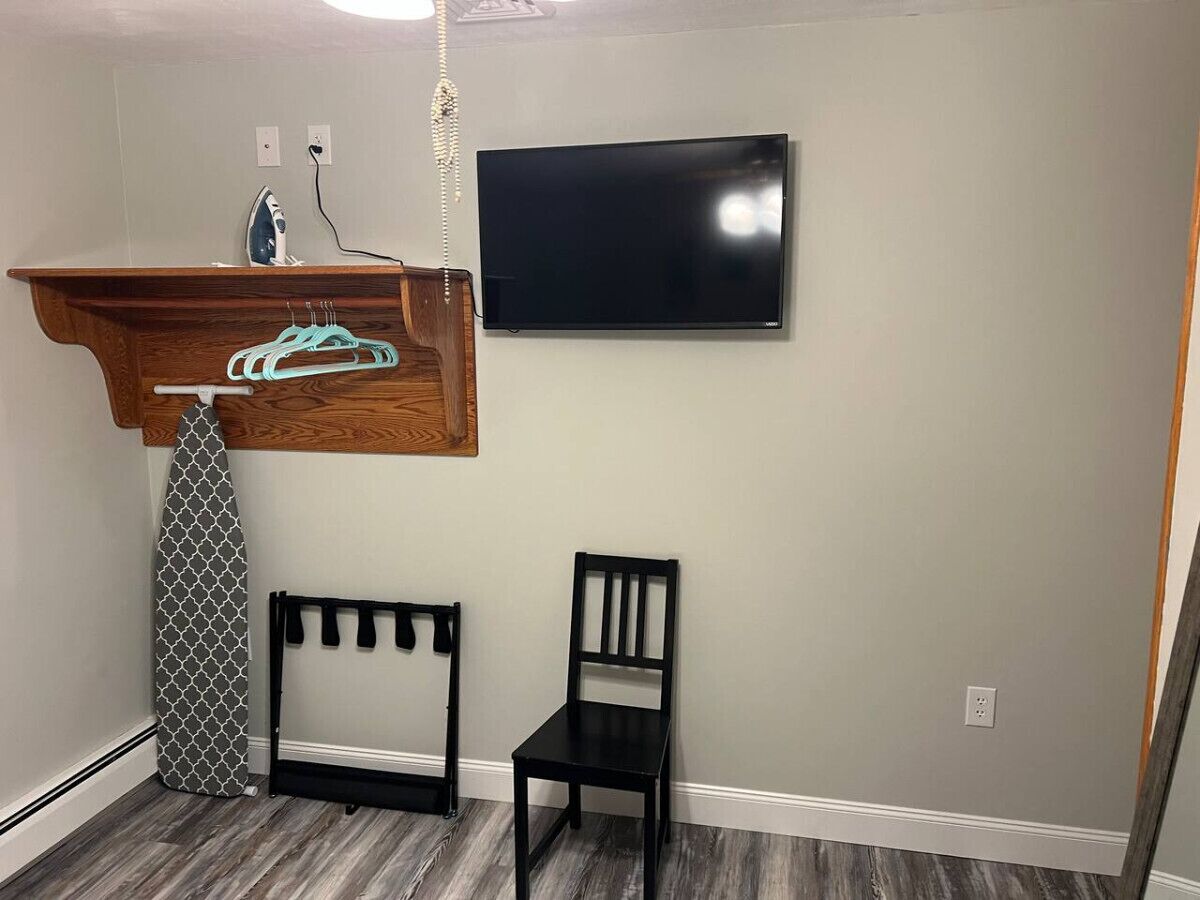 Een kamer met een houten plank waarop een strijkijzer en een kledingstuk liggen, een zwarte stoel en een flatscreen televisie aan de muur.