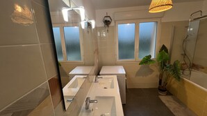 Ein modernes Badezimmer mit einer großzügigen Badewanne, zwei Waschbecken, Milchglasfenstern und einer Topfpflanze.