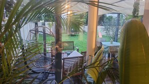 Ein überdachter Außenbereich mit Tisch und Stühlen, umgeben von viel Grün und Blick auf eine Rasenfläche.