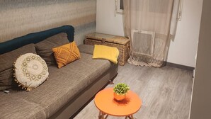 Ein Wohnzimmer mit einer Couch, Kissen, einem kleinen Tisch mit einer Pflanze, einem Wandzeichen und einem Fenster mit Vorhängen.