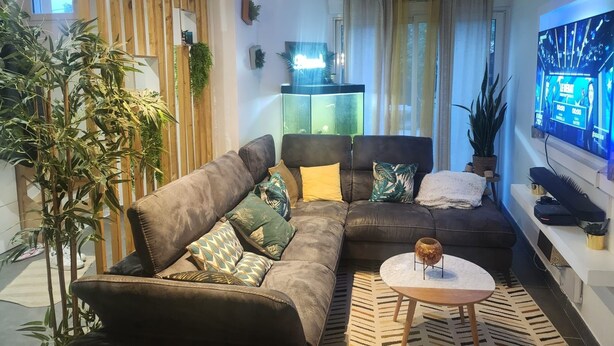 Ein modernes Wohnzimmer mit einer Eckcouch, einem Couchtisch und einem Fernseher. Pflanzen und ein dekoratives Regal sind ebenfalls vorhanden.