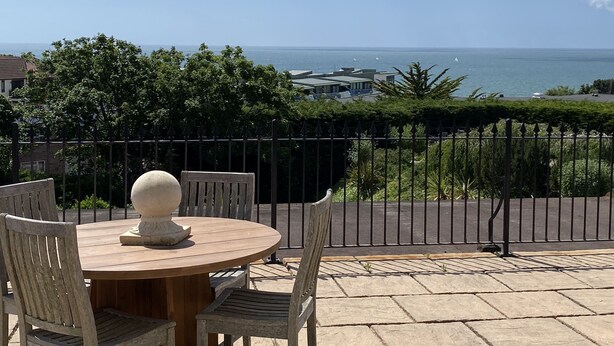 Una mesa redonda de madera con un objeto decorativo blanco, rodeada de sillas de madera, con vistas a la costa bajo un cielo despejado y una valla al frente.