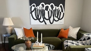 A living room with a green sofa, two orange pillows, a glass coffee table, and a large abstract painting on the wall. - Unique Design | Hot Tub | Brookside | Gathering Pl (Tulsa)
