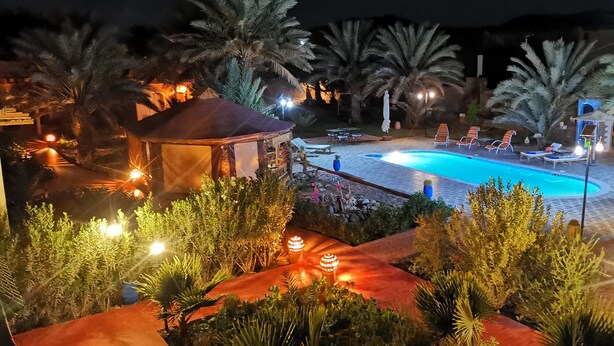 A nighttime pool area with lounge chairs, a gazebo, and palm trees. - Oasis Tilogui (Zagora)