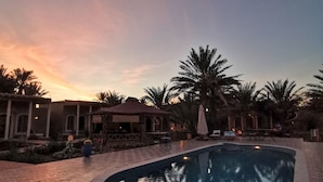 A pool area with a tiled deck, palm trees, and a building with arched windows. - Oasis Tilogui (Zagora)
