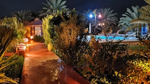 A nighttime view of a resort with a pool, a covered seating area, and a pathway lined with plants. - Oasis Tilogui (Zagora)