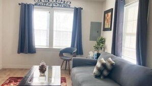 A living room with a grey sofa, a glass coffee table, a red patterned rug, a ceiling fan, and a window with blue curtains. - Cozy House (Dallas)