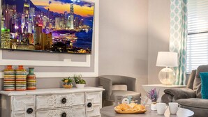 Ein Wohnzimmer mit Stadtbildgemälde, einer weißen Kommode mit gestreiften Vasen, einem runden Couchtisch mit Gebäck und Tassen sowie einem grauen Sofa.