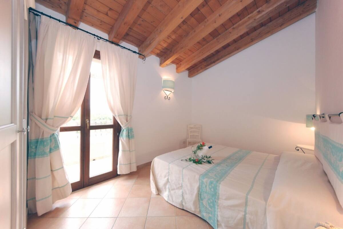 A bedroom with a bed, a window with curtains, a wall-mounted light, and a wooden ceiling.