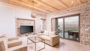 A modern living room with a stone wall, a flat-screen TV, a beige sofa, a glass coffee table, and a large sliding glass door leading to an outdoor area. - Triades Villas 3, Traumhafte Natursteinvilla mit Salzwasserpool, Ruhige Umgebung (Sivas)