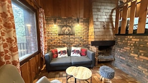 L’intérieur d’un chalet accueillant, avec une cheminée, un canapé agrémenté de coussins à motifs d’ours, une table ronde et un banc en bois.