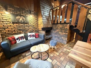 A cozy living area with a brick fireplace, a sofa with bear-themed cushions, and a small round table. - LA POMME DE PIN 2  Appartement avec terrasse (La Bresse)