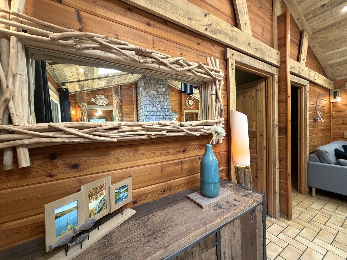 A rustic wooden interior with a wooden bench, a mirror with a woven frame, a vase, and framed pictures on the wall.