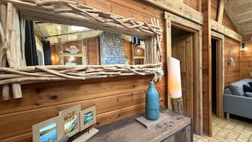 A rustic wooden interior with a wooden bench, a mirror with a woven frame, a vase, and framed pictures on the wall.