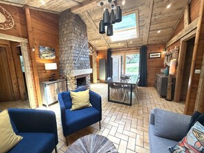 A log cabin interior with a stone fireplace, blue sofas, a dining area, and a large window.