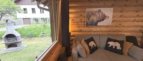 A cozy cabin interior with a wooden ceiling, a stone fireplace, a gray sofa with bear-themed pillows, and a painting of a bear on the wall.