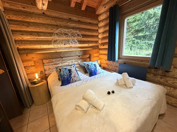A log cabin bedroom with a bed, pillows, a nightstand, and a window with a view of trees.