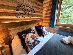 A wooden cabin-style room with a bed, two pillows with colorful dog faces, a geometric wall decoration, and a window with a forest view.