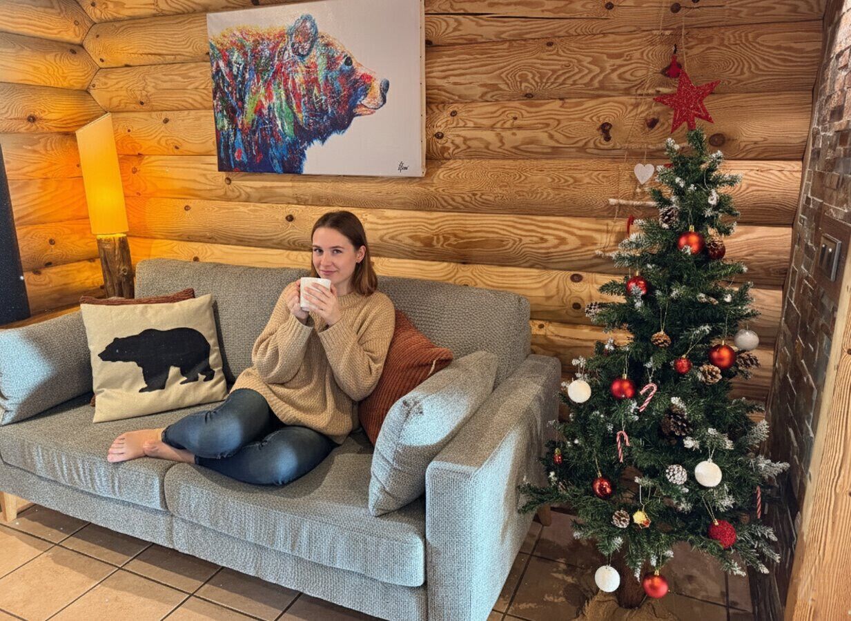 A person sitting on a grey sofa with a bear pillow, a colorful abstract painting on the wall, and a decorated Christmas tree.