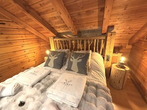 Un dormitorio de cabaña de madera con una cama, dos almohadas con diseños de ciervo, una manta blanca y esponjosa y una mesita de noche de madera con una lámpara.
