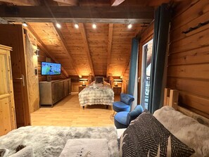 Interior de una cabaña de troncos con techo de madera, una cama con una manta de piel, un sillón azul y un televisor sobre un mueble de madera.