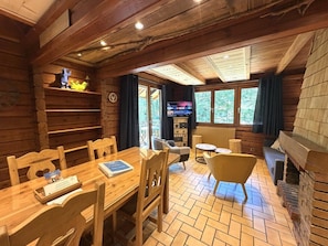 Ein Holzhütten-Innenraum mit Esstisch, Stühlen, Kamin, Fernseher und einem Fenster mit Blick auf Bäume.