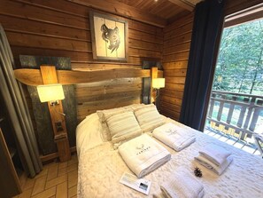 Quarto de cabana de madeira com uma cama, duas luminárias fixadas na parede, uma obra de arte com a cabeça de um animal emoldurada e uma janela com vista para árvores.