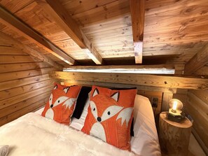 A wooden cabin interior with a bed, fox-themed pillows, a nightstand with a lamp, and a wooden ceiling.