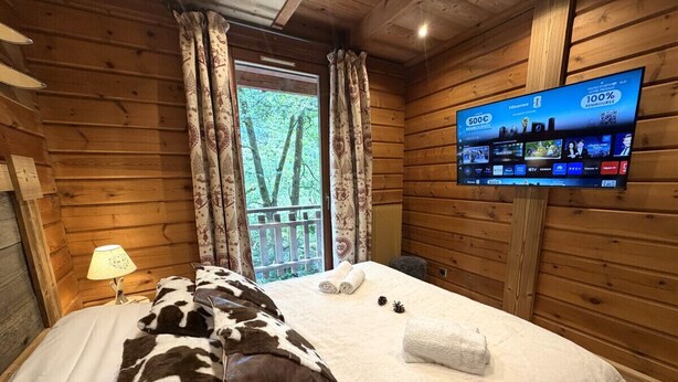 A wooden cabin room with a bed, a TV, and a window with curtains. - LE MONTAGNARD Chalet en bois (La Bresse)