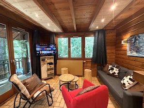 A wooden cabin interior with a sofa, chairs, a TV, and a window overlooking a forest.
