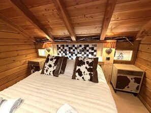 A wooden cabin bedroom with a bed, cow-patterned pillows, a checkered headboard, and two bedside tables with lamps.