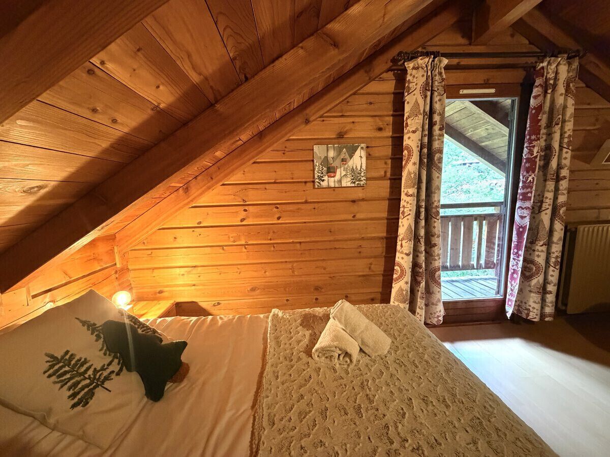 Un dormitorio acogedor en el ático con techo inclinado, cama con manta blanca y almohada, una ventana pequeña con cortinas, y un cuadro en la pared.