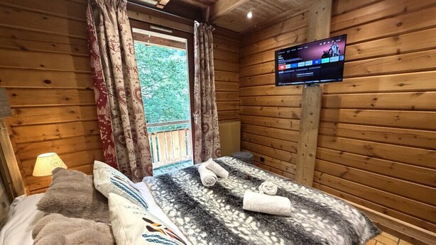A wooden cabin room with a bed, two pillows, a blanket, a TV on the wall, and a window with curtains.