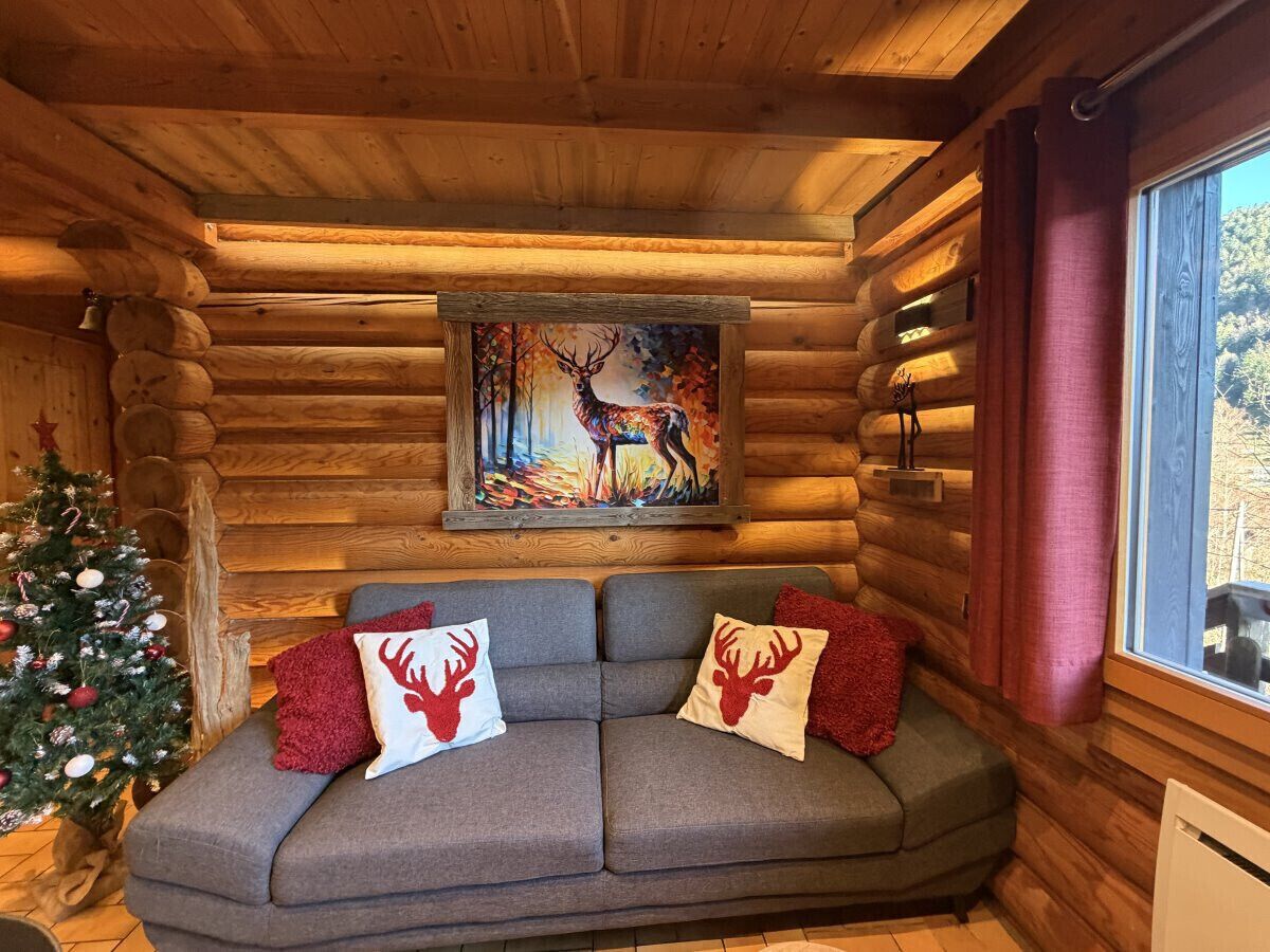 Interior acogedor de una cabaña de troncos, con un sofá decorado con almohadones con motivos de renos, un cuadro de un ciervo y un árbol de Navidad adornado.