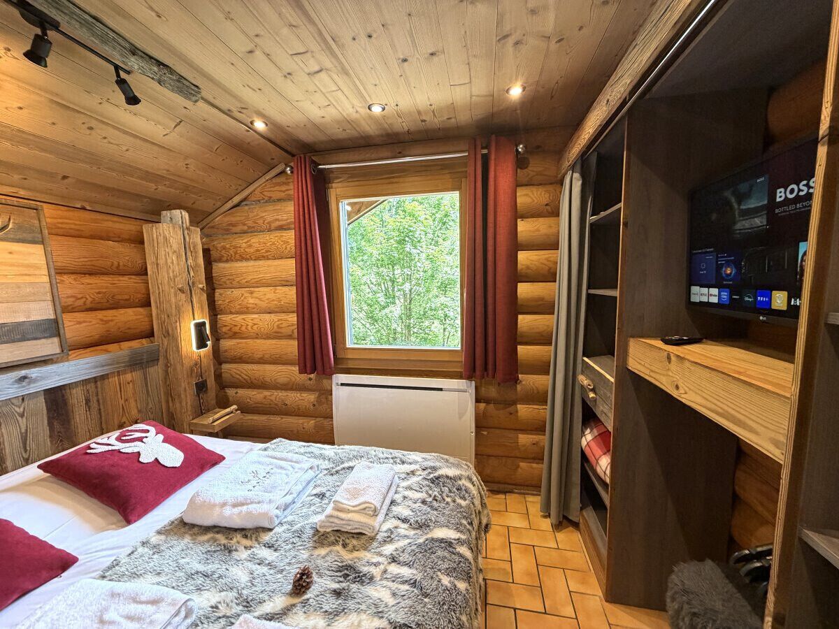 Une pièce de type cabane en bois, avec un lit, une fenêtre avec des rideaux, un radiateur et un téléviseur posé sur une étagère.