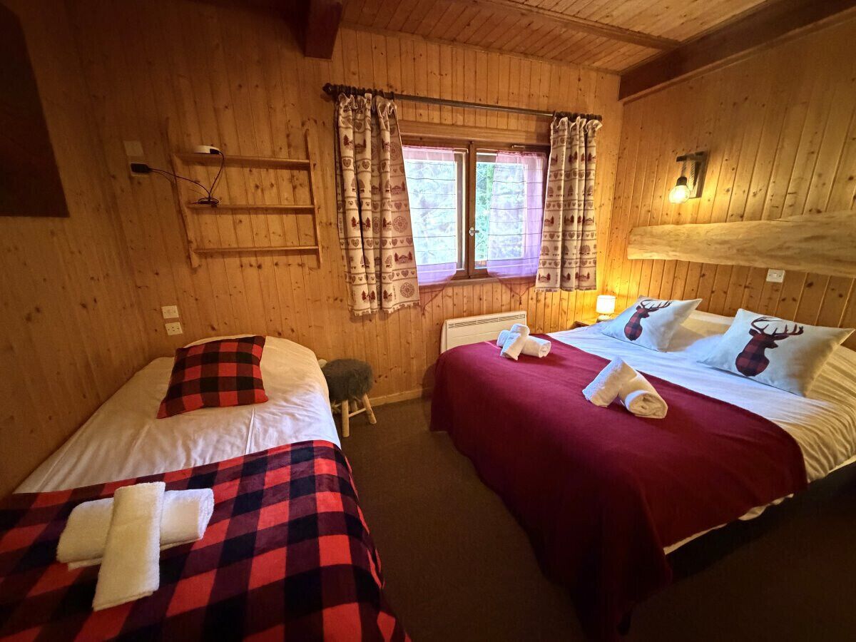 A wooden room with two beds, a window with curtains, a small stool, and a wall-mounted light fixture.