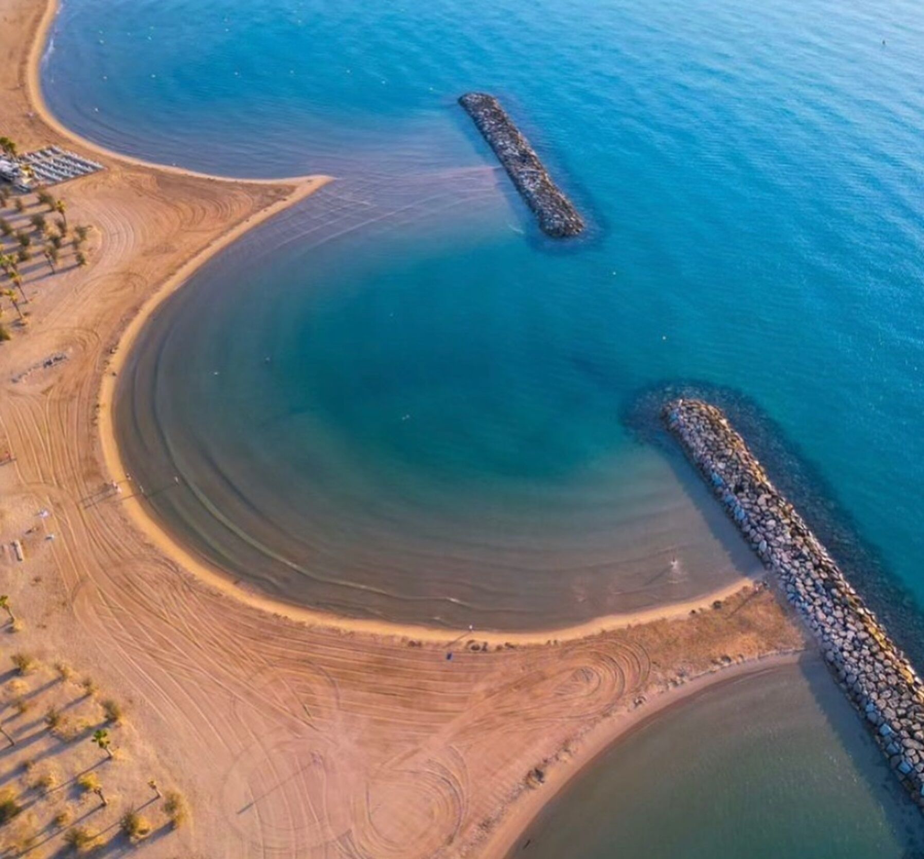An aerial view of a coastal area with a sandy beach, a rocky breakwater, and a clear blue sea.
