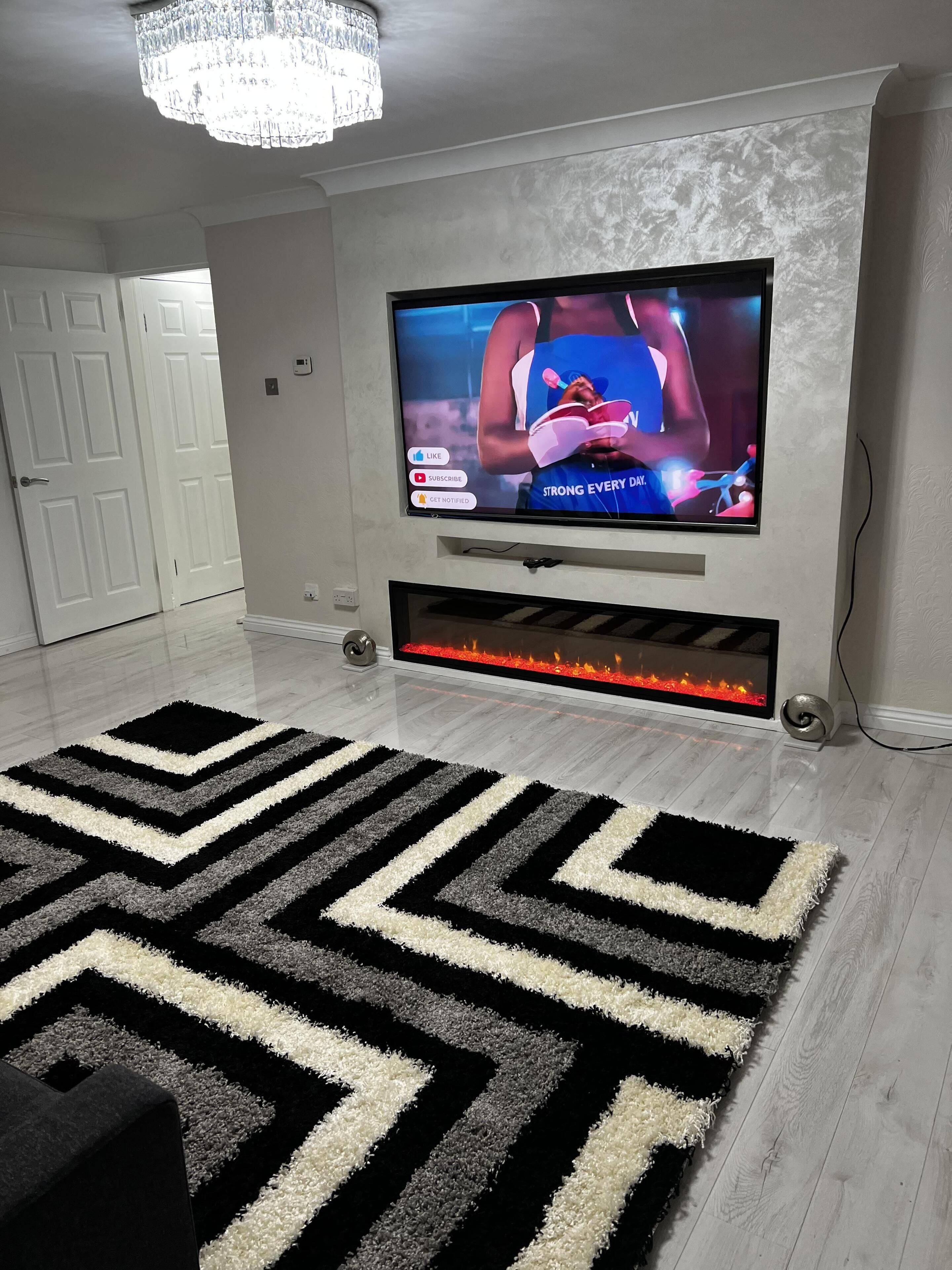 Ein modernes Wohnzimmer mit einem Flachbildfernseher an einer strukturierten Wand, einem schwarz-weißen Zickzack-Teppich und einem eingebauten Kamin mit Feuer.