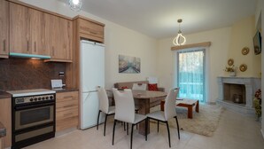 A modern kitchen with wooden cabinets, a built-in oven, and a refrigerator. There is a dining area with a table and chairs, a fireplace, and a window with curtains. - The Miller House under Meteora (Kalampaka)