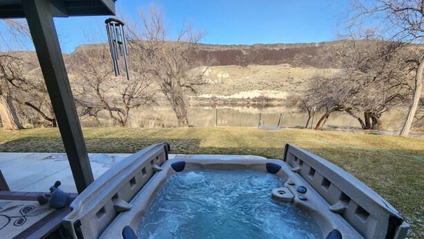 Un jacuzzi con revestimiento de piedra, rodeado de césped y vistas a un lago y colinas.