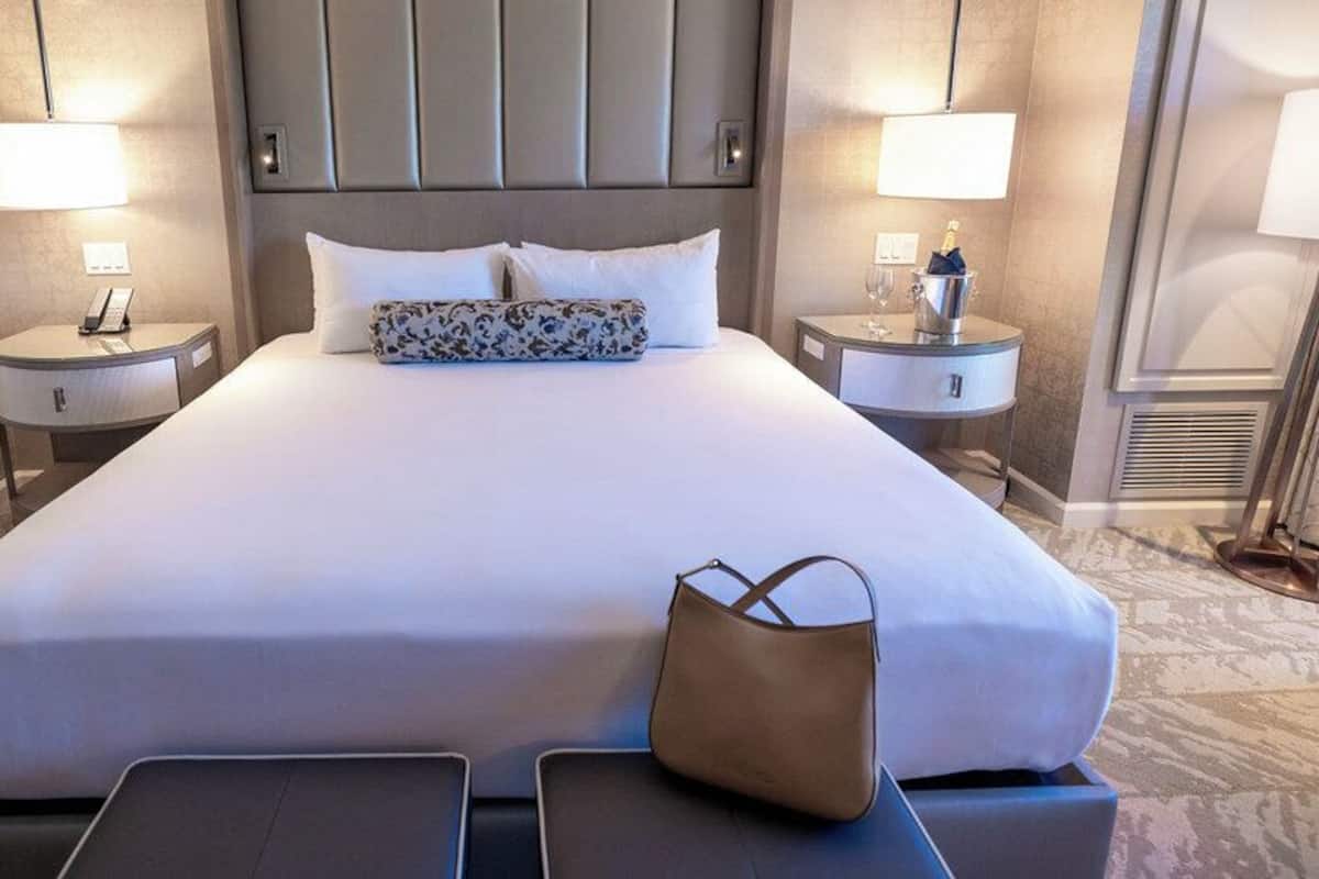 Una habitación de hotel con una cama grande, dos mesitas de noche, una lámpara, una botella de champán y un bolso sobre la cama.