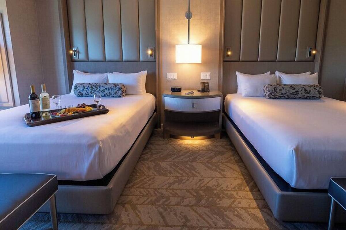 Habitación de hotel con dos camas, una mesita de noche con lámpara y un atril con botellas y vasos.