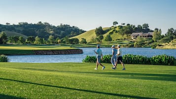 Un campo de golf con una masa de agua, colinas verdes y un edificio al fondo.