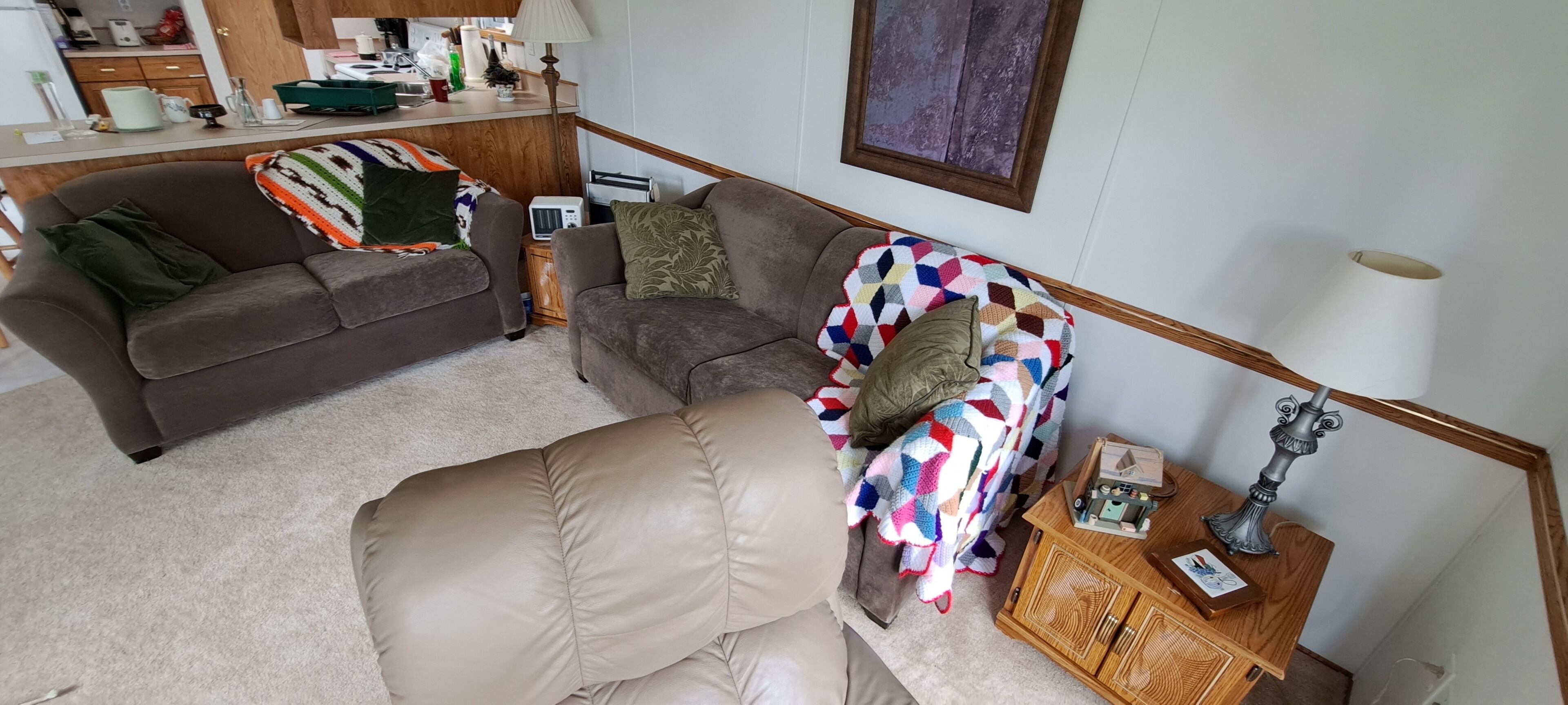 Ett mysigt vardagsrum med en brun soffa, en beige fåtölj och en färgglad pläd. Där finns ett träskåp med en lampa och ett litet bord med en fotoram.