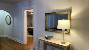 En hall med trägolv, en ljusblå vägg, en spegel med guldfärgad ram och ett bord med en lampa och en dekorativ skål.