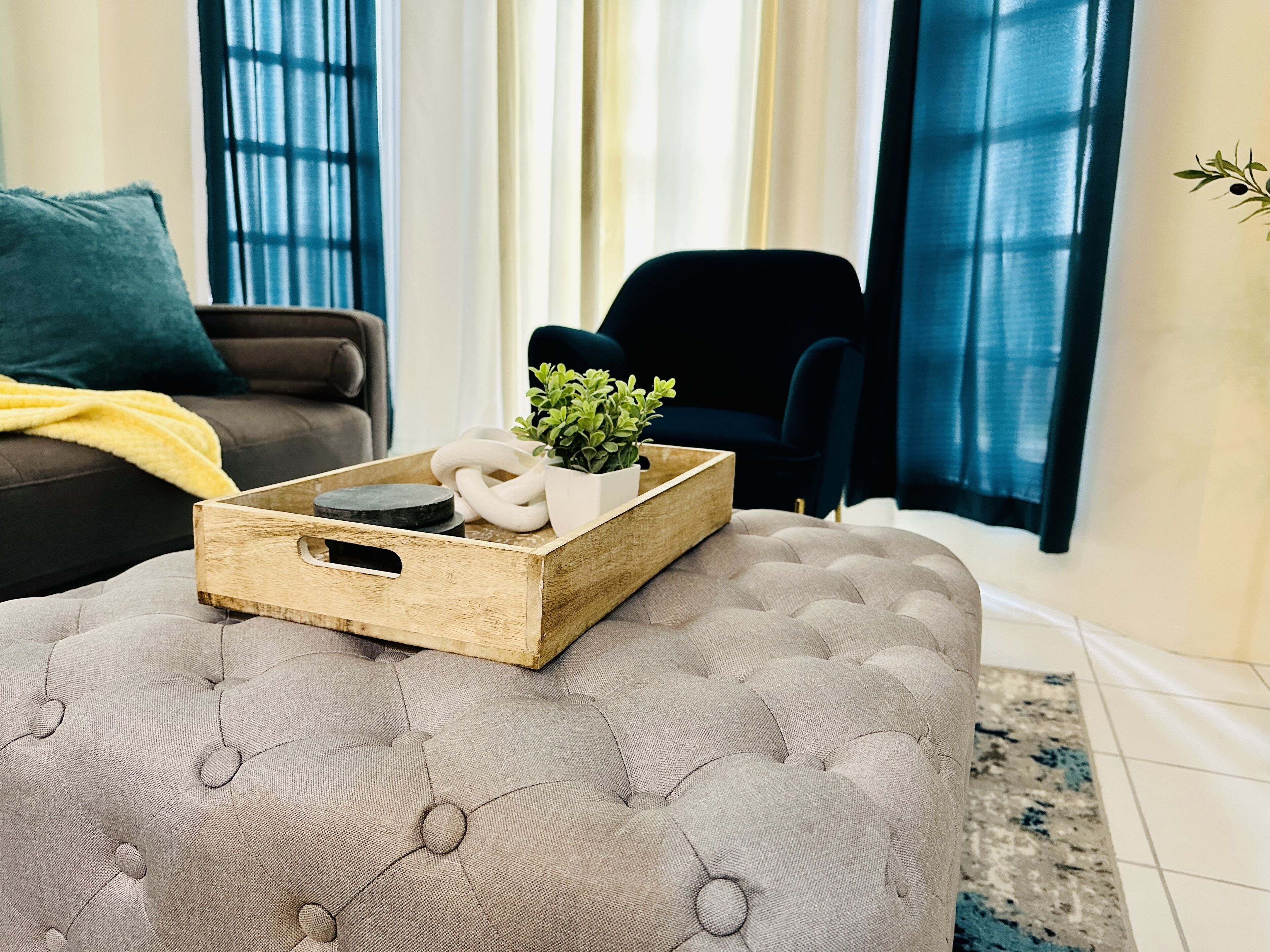 A living room with a grey tufted ottoman, a wooden tray with decorative items, a dark blue armchair, and a sofa with a yellow throw blanket.