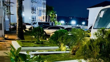 A nighttime view of a hotel with a well-lit building, a parking area, and a landscaped garden with palm trees and shrubs.