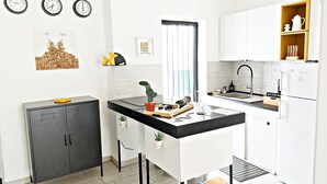A modern kitchen with white cabinets, a black countertop, and a small island. There are four clocks on the wall, a potted plant on the island, and a window with blinds. - Aurelio's Artpartment - "Unconventional House" (Siracusa)