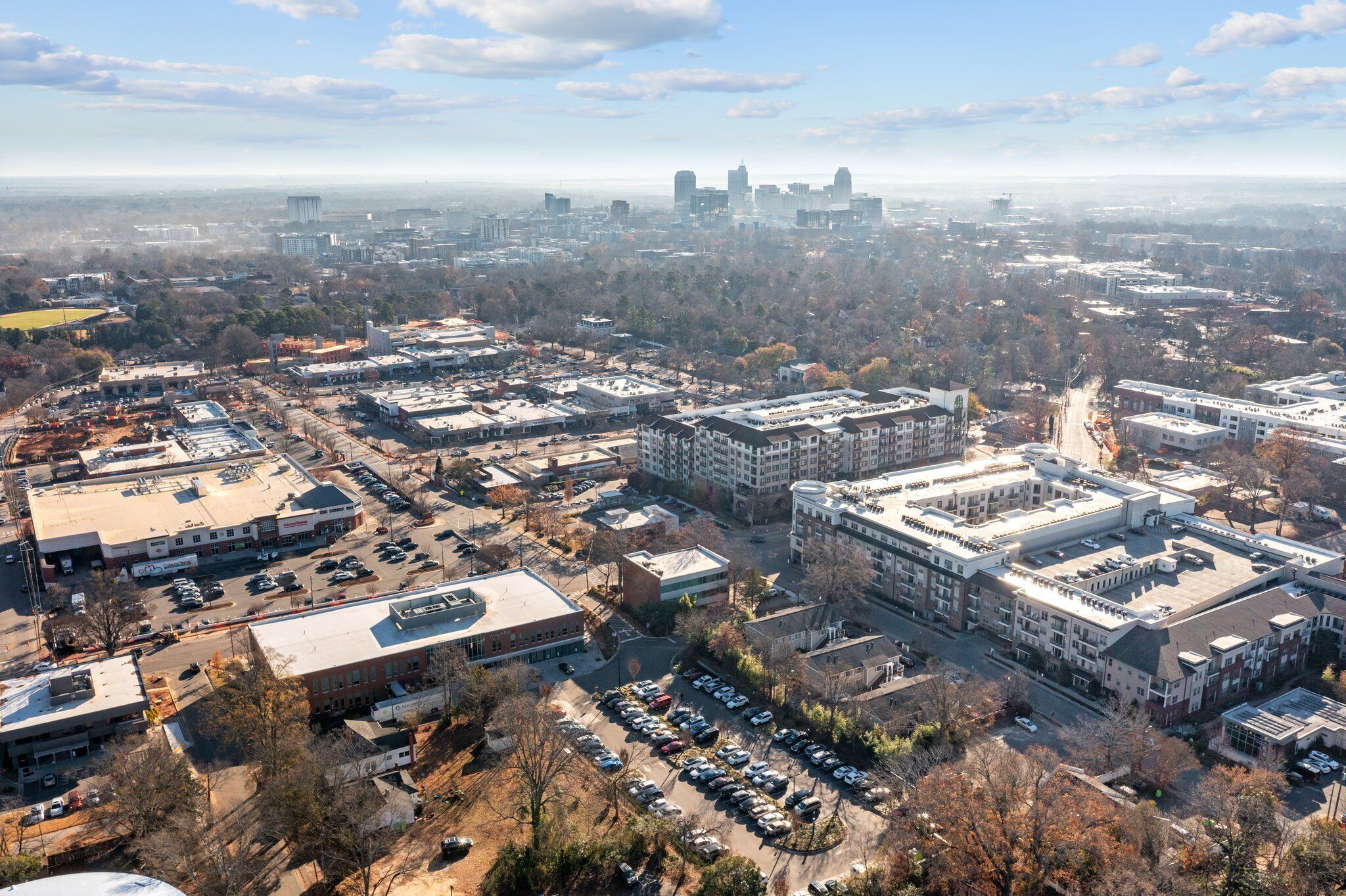 An aerial view of a cityscape with a mix of commercial and residential buildings, a large parking lot, and a distant skyline.