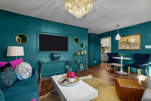 A teal-themed living room with a flat-screen TV, a coffee table with decorative items, a sofa with cushions, and a dining area with chairs and a table. - The Jewel Box- NC State/ Walkable (Raleigh)